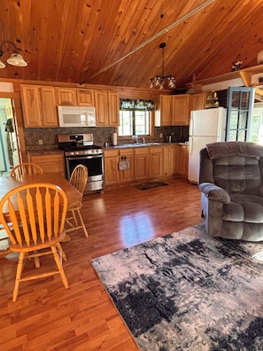 A wooden cabin interior with a kitchen, dining area, and a plush armchair. - Little lake house, quiet cozy  (West Winfield)