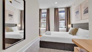 Habitación de hotel con dos camas, un espejo grande, un escritorio de madera y vistas a un edificio a través de las ventanas.