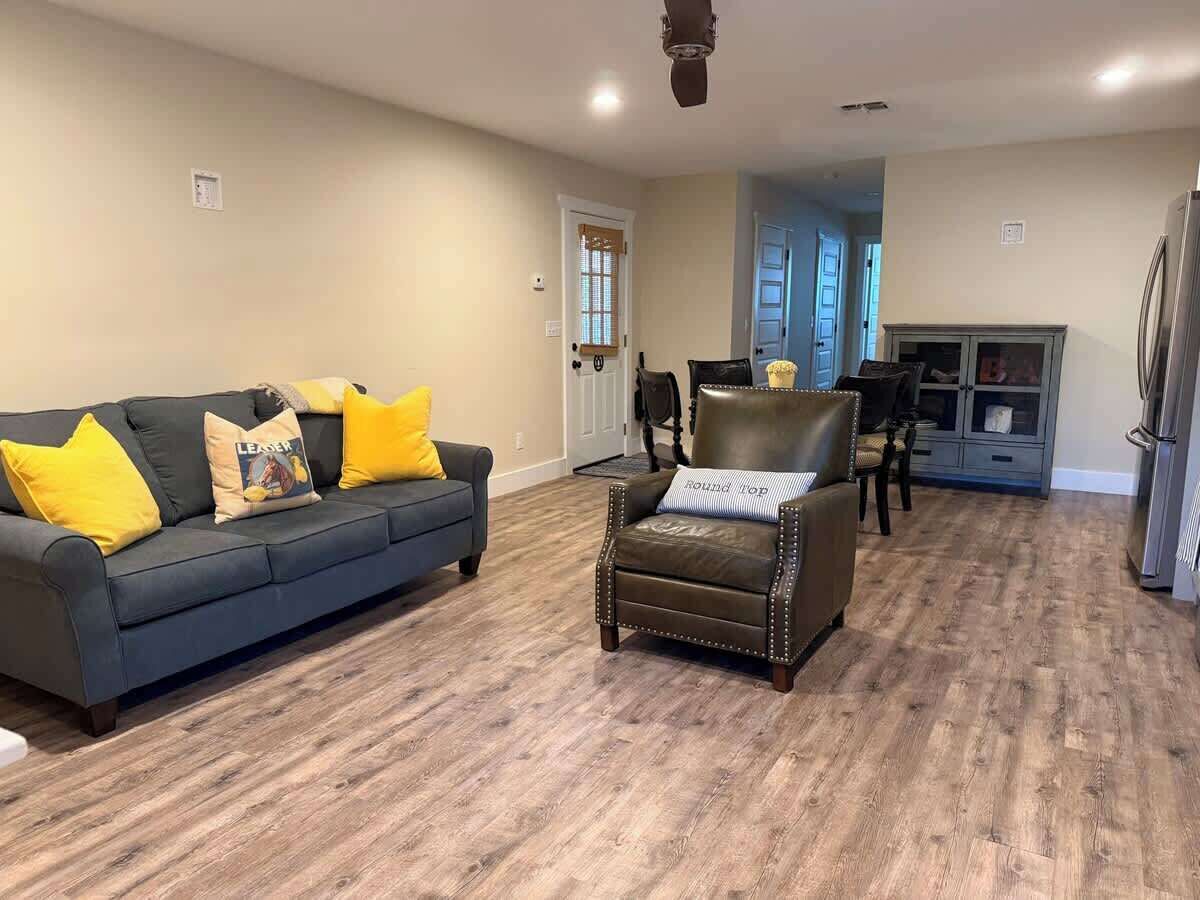 Una sala de estar con un sofá gris oscuro, cojines amarillos, un sillón de cuero marrón y piso de madera.