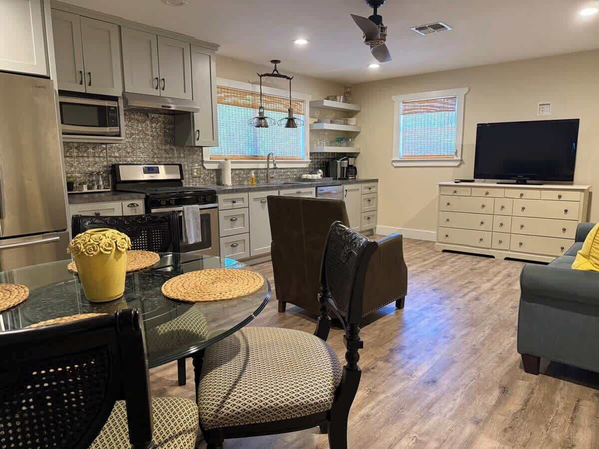 A modern kitchen with stainless steel appliances, a dining area with a glass table and chairs, and a living room with a sofa and a flat-screen TV.