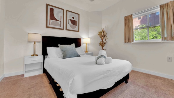 A neatly made bed with white linens, two bedside lamps, a nightstand, a decorative plant, and a window with curtains.