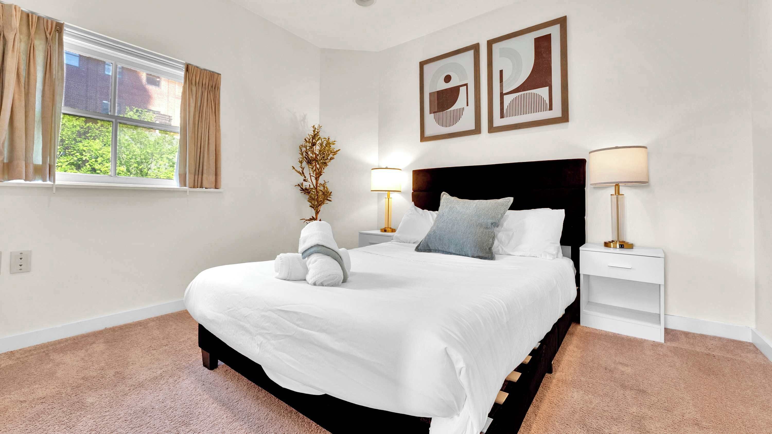 A hotel room with a large bed, two bedside tables with lamps, a window with curtains, and two framed artworks on the wall.