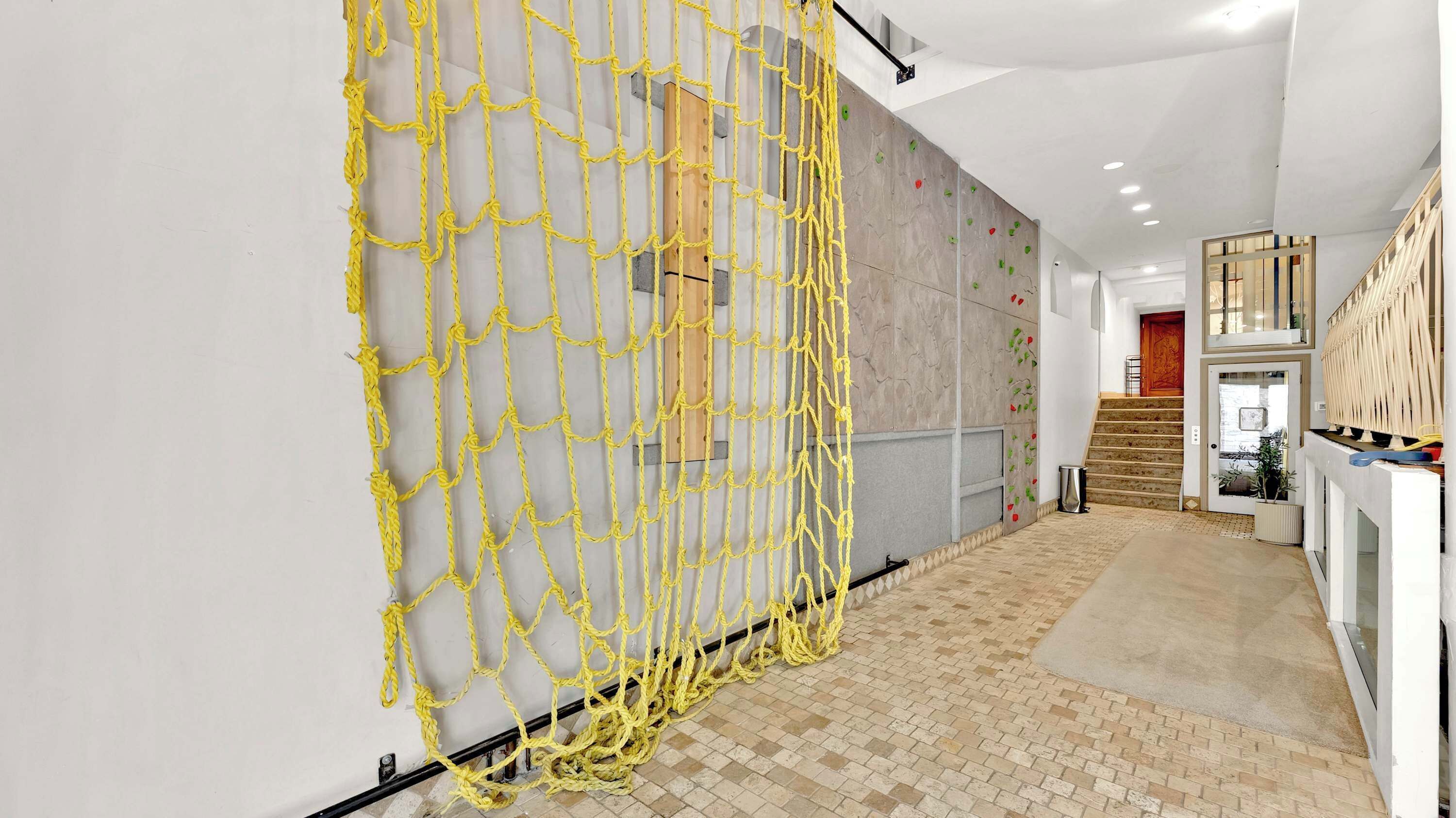 Un couloir avec une barrière faite d’une corde jaune, un escalier et un mur équipé de prises d’escalade.