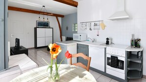 Une cuisine avec des meubles blancs, un four à micro-ondes, un four et un évier. On y trouve une chaise et une table sur laquelle est posé un vase de fleurs.