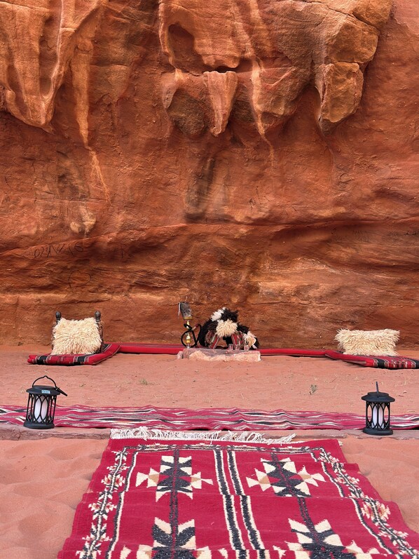 A traditional setting with a red carpet, cushions, and a hookah, set against a red rock wall.