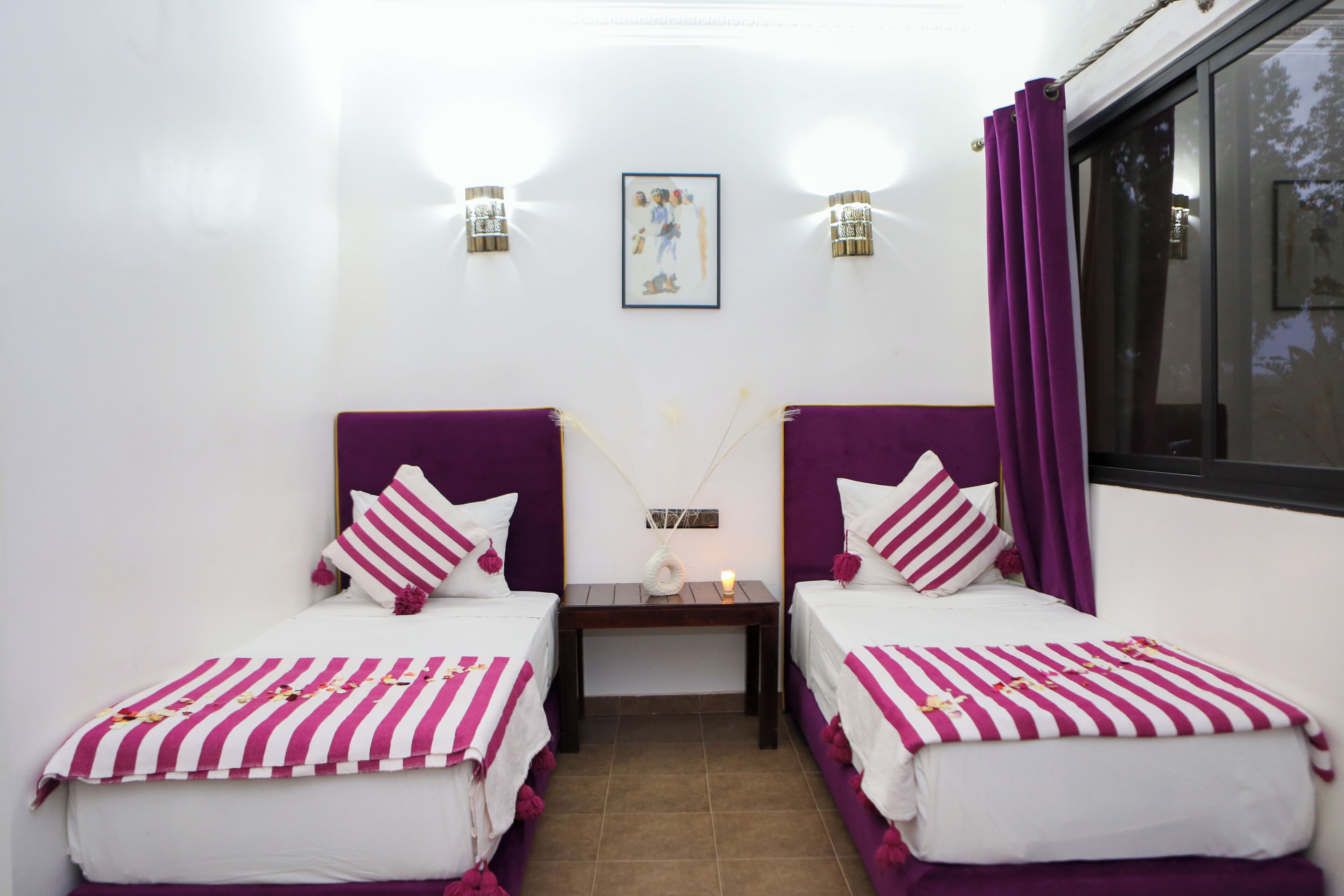 Ein Doppelzimmer mit zwei Betten, jeweils mit violettem Kopfteil und weißer Bettwäsche mit pinken Streifen. Zwei Wandlampen und ein gerahmtes Bild sind über den Betten angebracht.