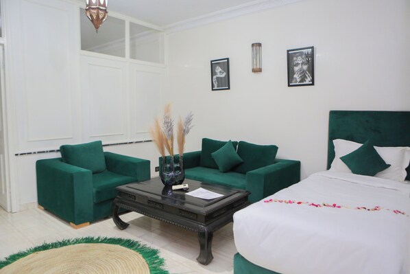 A hotel room with a green sofa set, a wooden coffee table, a white bed with green pillows, and two framed pictures on the wall.