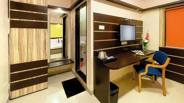 A modern hotel room with a wooden accent wall, a desk with a chair, a television, and a small table with a vase of flowers. - Hotel Golden Treat (Indore)