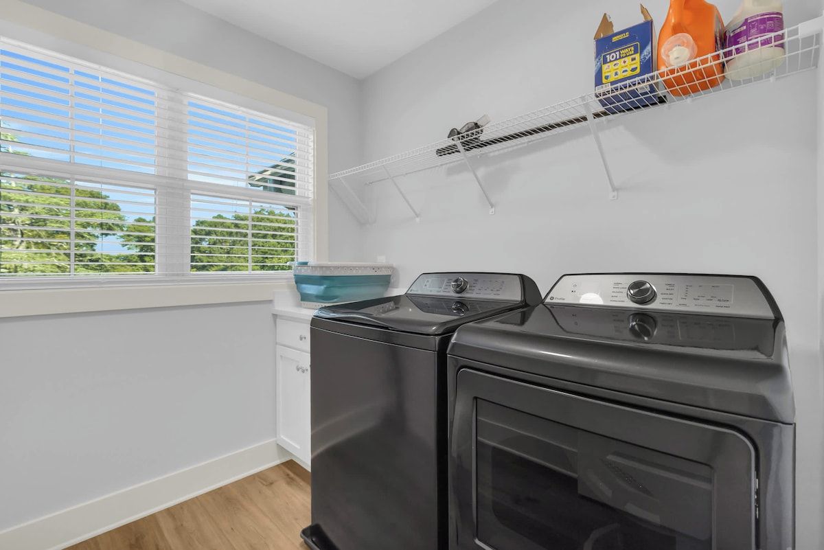 A laundry room with two washing machines, a window with blinds, and a shelf with cleaning supplies.