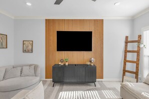 Un salon avec un téléviseur à écran plat fixé sur un mur recouvert de panneaux en bois, une console de couleur foncée agrémentée d’objets décoratifs, un canapé de couleur claire et une échelle en bois appuyée contre le mur.