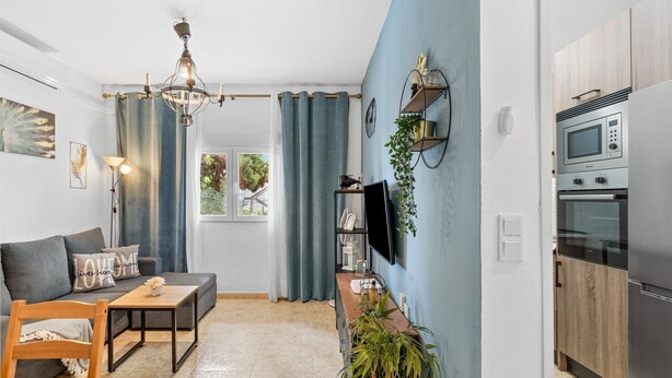 Ein modernes Wohnzimmer mit Sofa, Esstisch, einer Chandelierleuchtre, einem an der Wand montierten Fernseher und einer Küche mit Backofen und Mikrowelle.