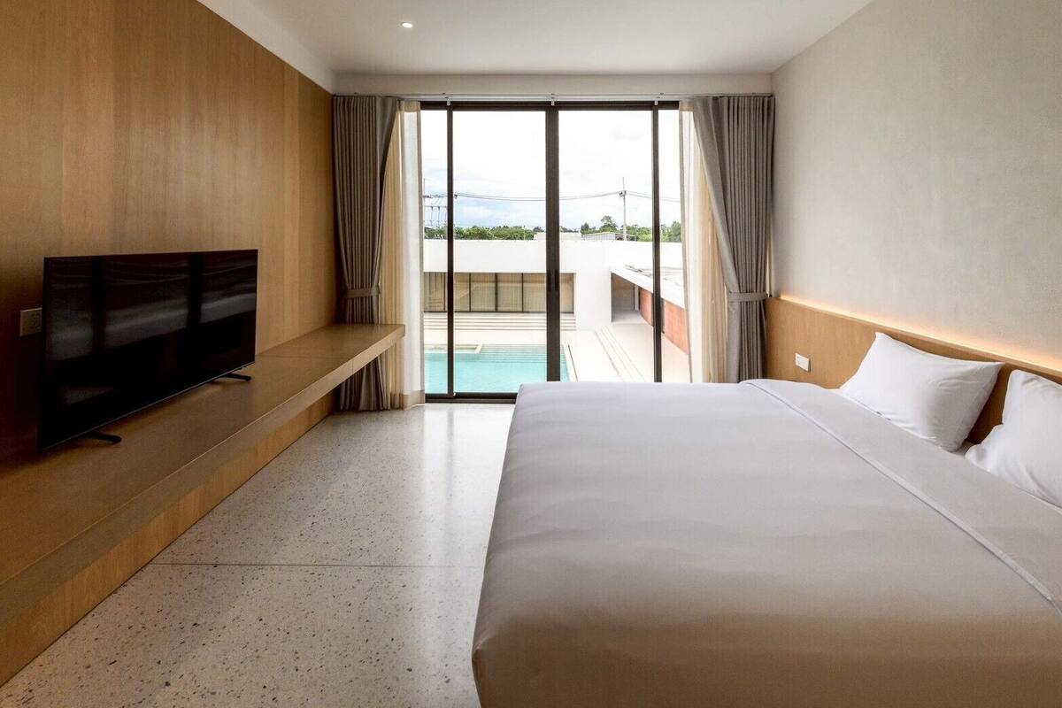 A hotel room with a large bed, a flat-screen TV on a wooden shelf, and a view of a swimming pool through a glass door.