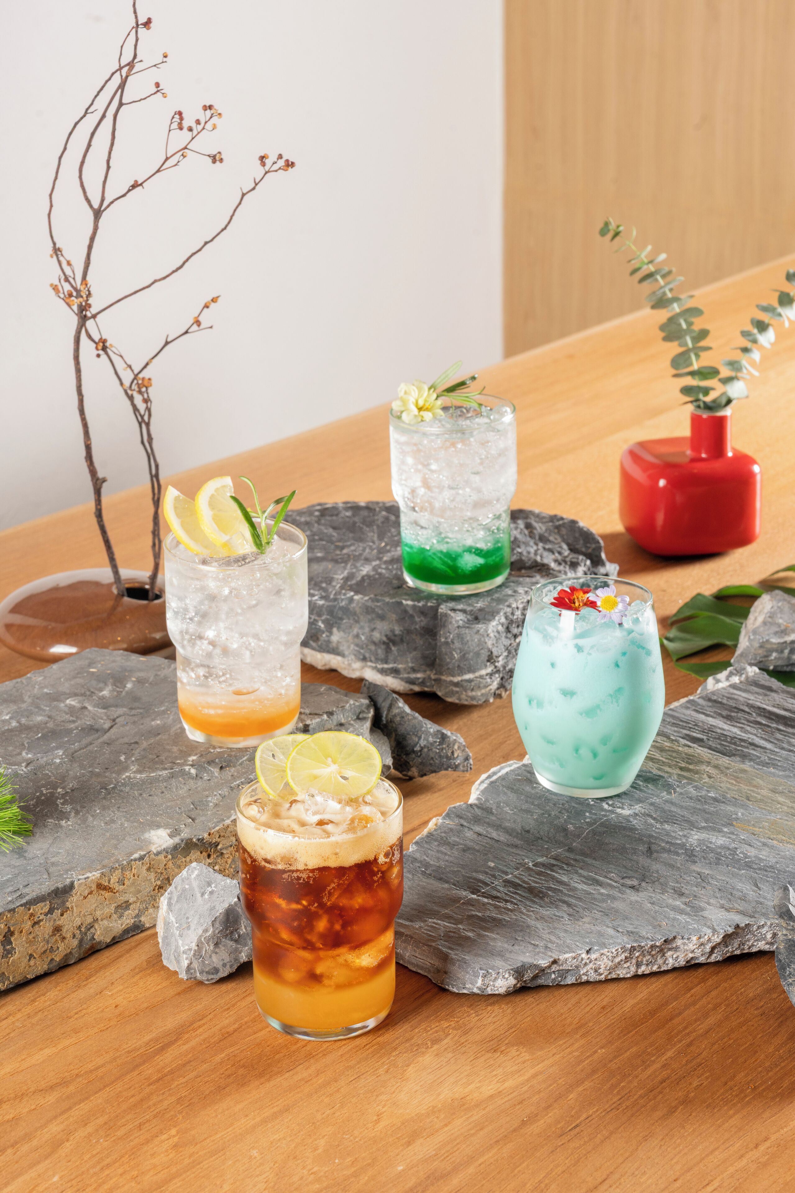 Cuatro vasos de bebidas heladas sobre una mesa de madera, con pedazos de hielo y una planta pequeña.
