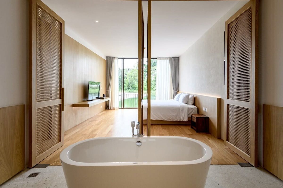 A modern bathroom with a freestanding bathtub, a vanity with a mirror, and a bed with a view of the outdoors.