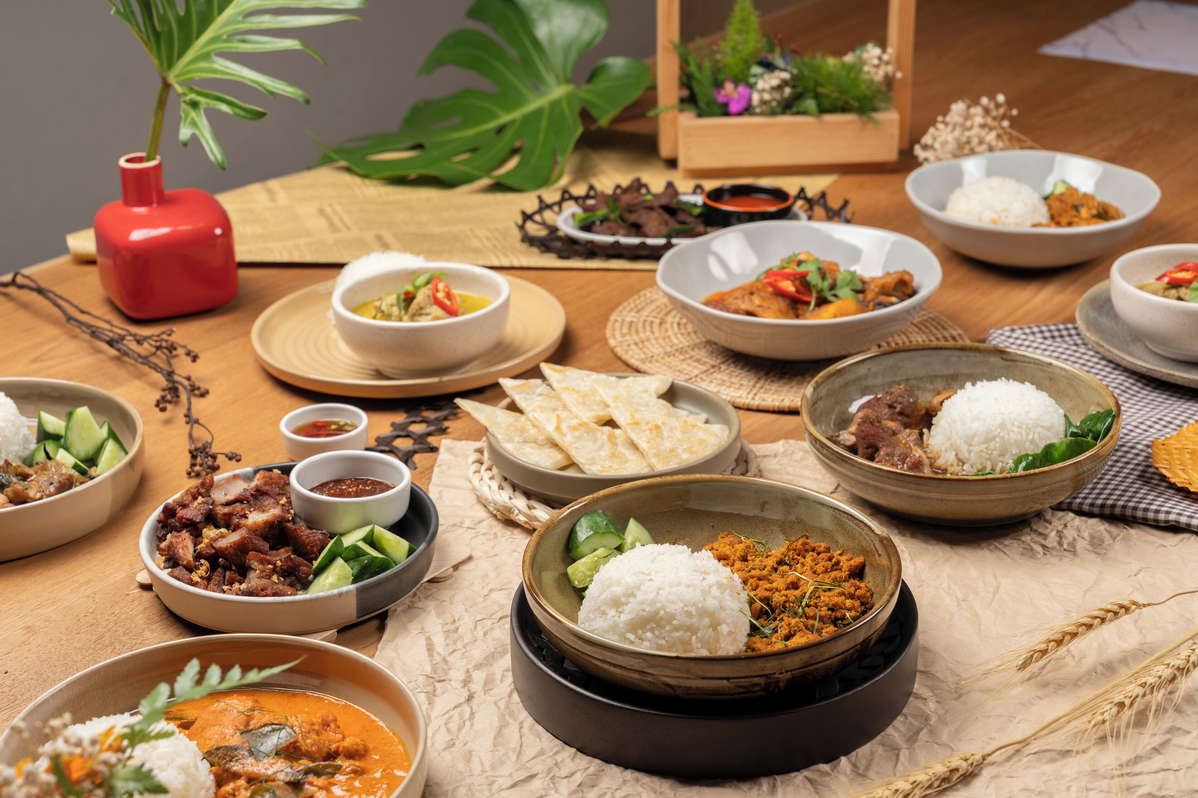 A variety of dishes including rice, meat, and vegetables, arranged on a wooden table with a red vase and a plant in the background.