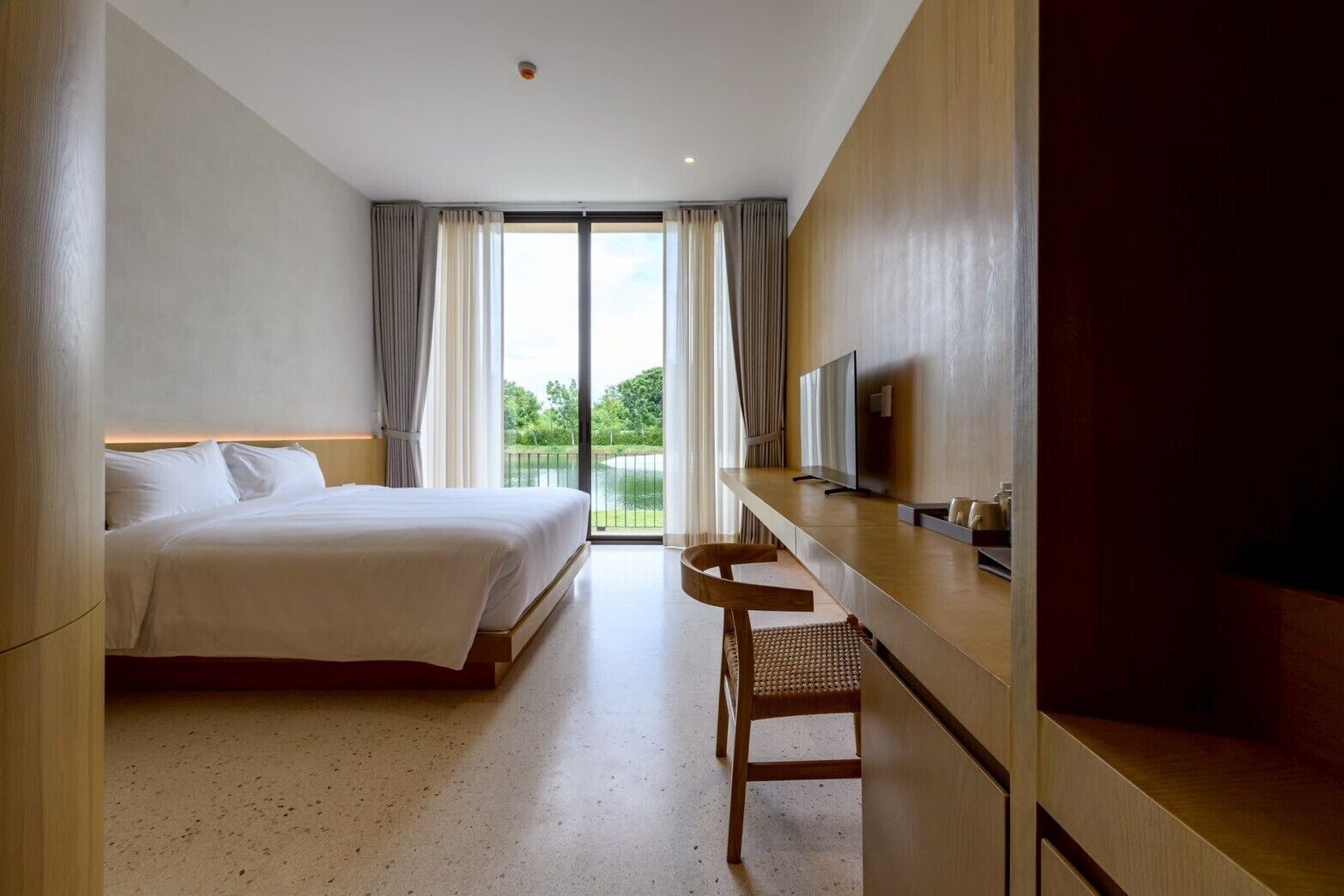 A hotel room with a bed, a desk with a chair, a TV, and a view of greenery through the window.