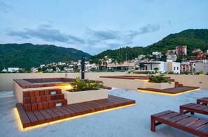 A rooftop terrace with wooden steps and seating areas, overlooking a cityscape with buildings and mountains.