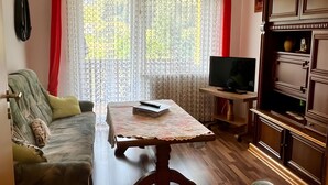 Een woonkamer met een houten vloer, een tafel met een kantkleed, een bank, een televisie en een kast.