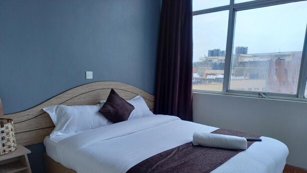 Una cama bien tendida con ropa de cama blanca y marrón oscuro, un cabecero de madera y vistas a edificios a través de una ventana.
