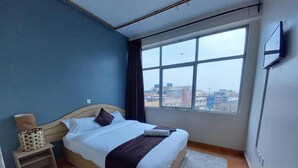 Habitación de hotel con una cama grande, una mesita de noche pequeña, una silla, un televisor montado en la pared y una ventana con vista a la ciudad.