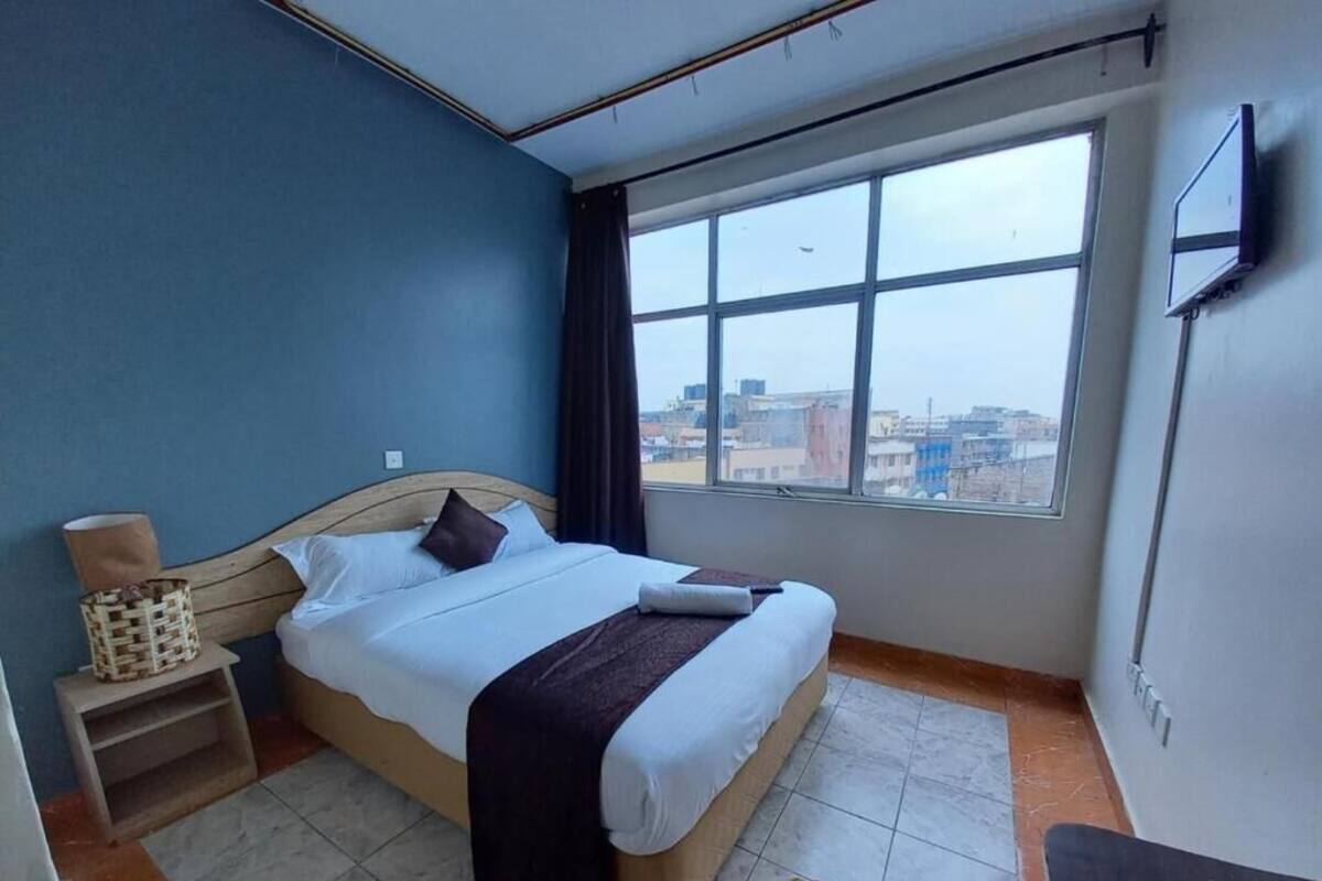 A hotel room with a large bed, a small bedside table, a chair, a TV mounted on the wall, and a window with a city view.