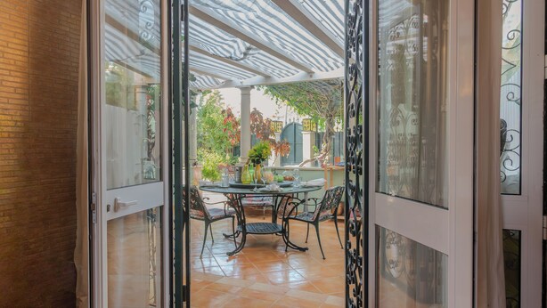 Eine offene Glasdoor führt zu einer Terrasse mit Tisch und Stühlen, umgeben von Pflanzen und einem gefliesten Boden.