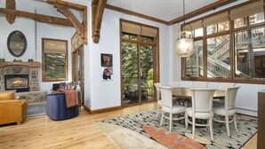 A cozy living room with a fireplace, wooden beams, a dining area with a table and chairs, and a large window offering a view of the outdoors.