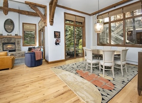 Uma sala de estar aconchegante com lareira, vigas de madeira, área de jantar com mesa e cadeiras, e uma janela grande com vista para o exterior.