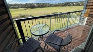 A balcony with a glass table and two chairs, overlooking a grassy field and buildings in the distance. - Coastal Cove Suite with balcony - close to hot springs. (Fingal)
