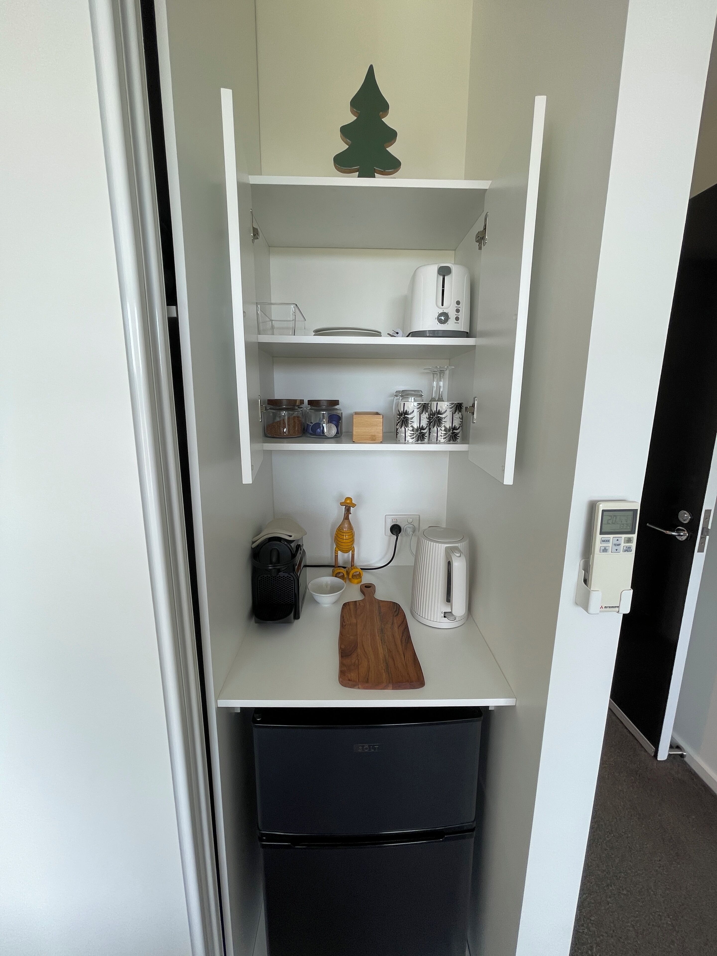 A compact kitchenette with a white countertop, a small black refrigerator, a wooden cutting board, a coffee maker, and a scale.