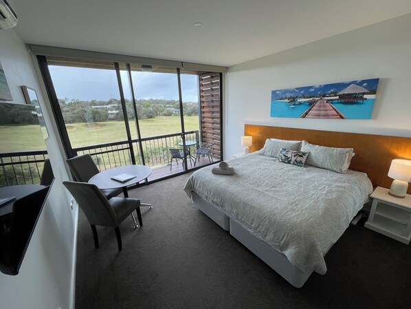 A hotel room with a large balcony, a bed with a patterned bedspread, a painting of a tropical scene, and a desk with a chair. - Coastal Cove Suite with balcony - close to hot springs. (Fingal)