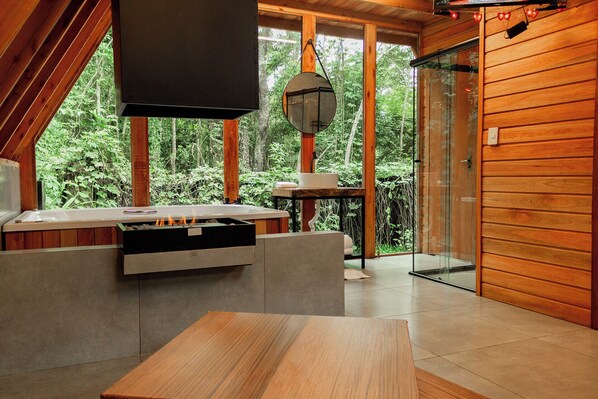 Une cabane moderne en bois, dotée d’une cheminée en pierre, d’une porte vitrée donnant sur un balcon et d’une grande fenêtre offrant une vue sur une forêt.