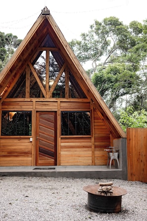 A wooden cabin with a triangular roof, large windows, and a fire pit in the foreground.