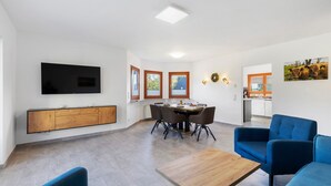 Un salon moderne doté d’un téléviseur à écran plat, d’un coin repas avec une table et des chaises, et d’une cuisine en arrière-plan.