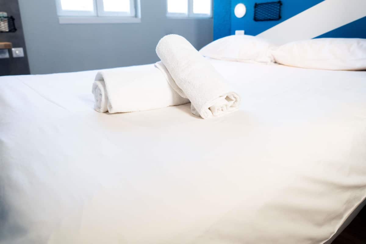 Ein ordentlich bezogenes Bett mit weißen Bettwäsche und einem zusammengerollten Handtuch, eine blaue Akzentwand und ein Fenster mit Jalousien.