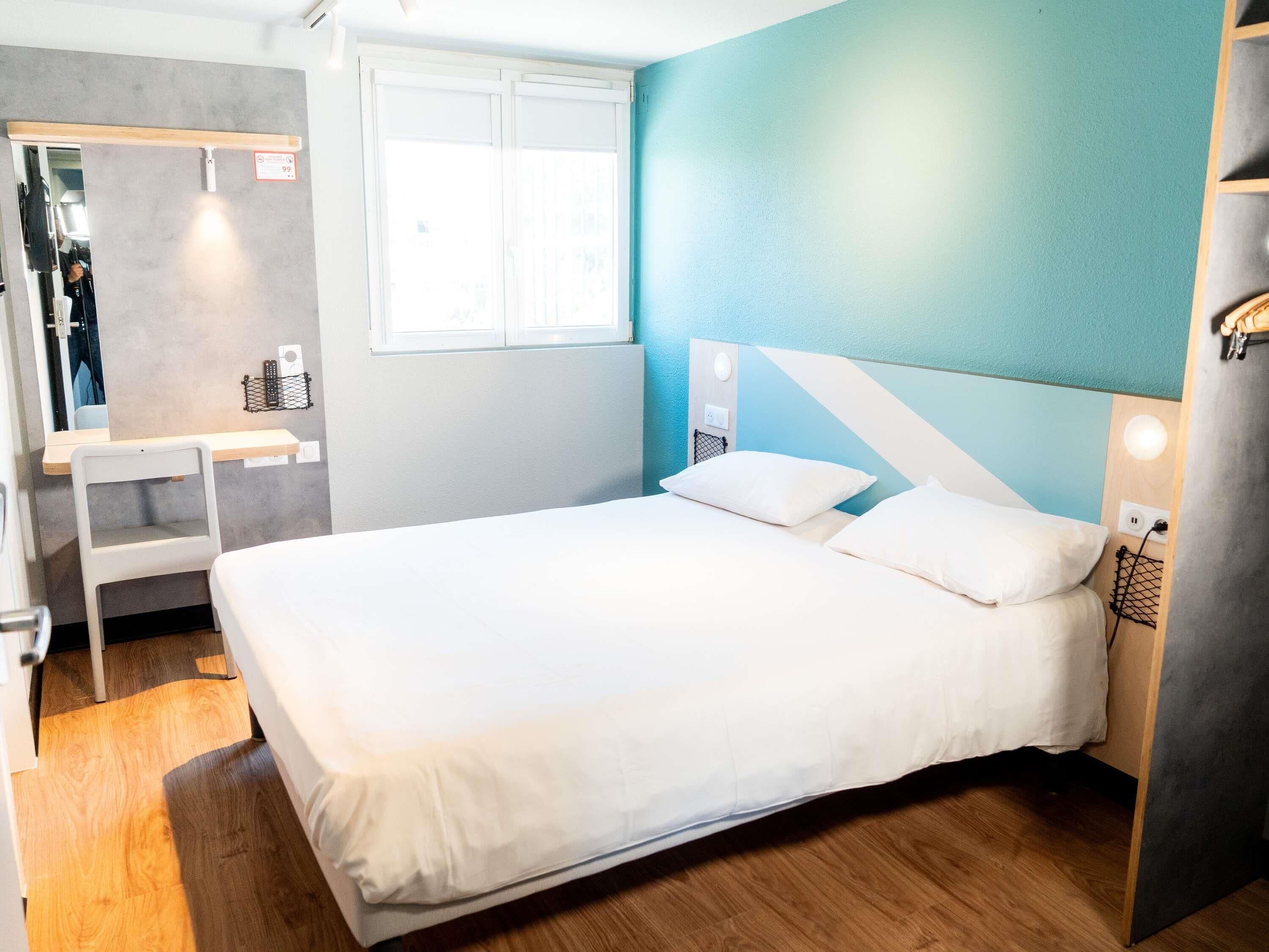 A bedroom with a bed, a desk, a mirror, and a window.