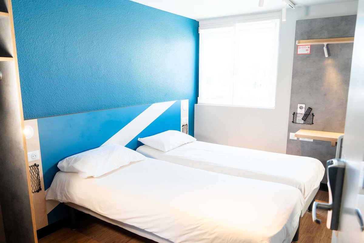 Ein Hotelzimmer mit zwei Betten, einer blauen Akzentwand, einem Fenster, einem Schreibtisch und einem kleinen Regal mit einer Fernbedienung.