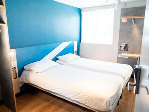 Habitación de hotel con dos camas, una pared de acento azul, una ventana, un escritorio y una pequeña estantería con un control remoto.
