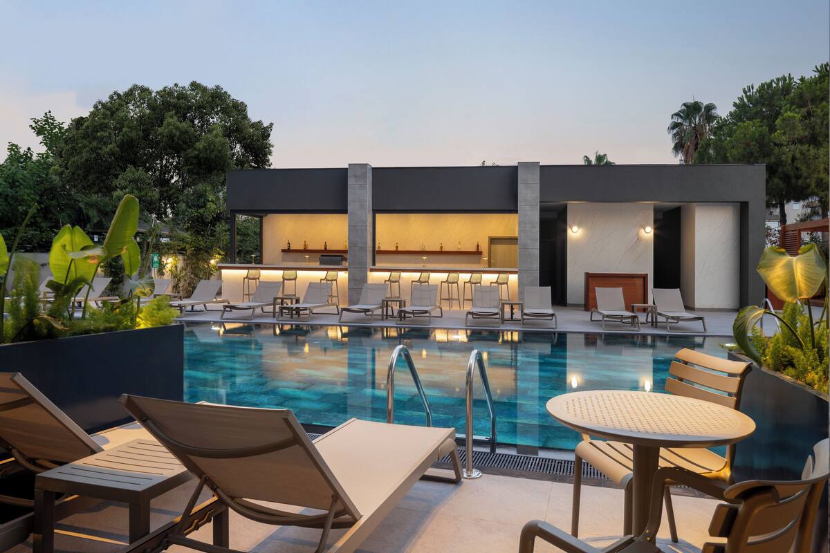 Un área de piscina moderna con sillones de descanso y una mesa, rodeada de follaje y un edificio con fachada de vidrio.