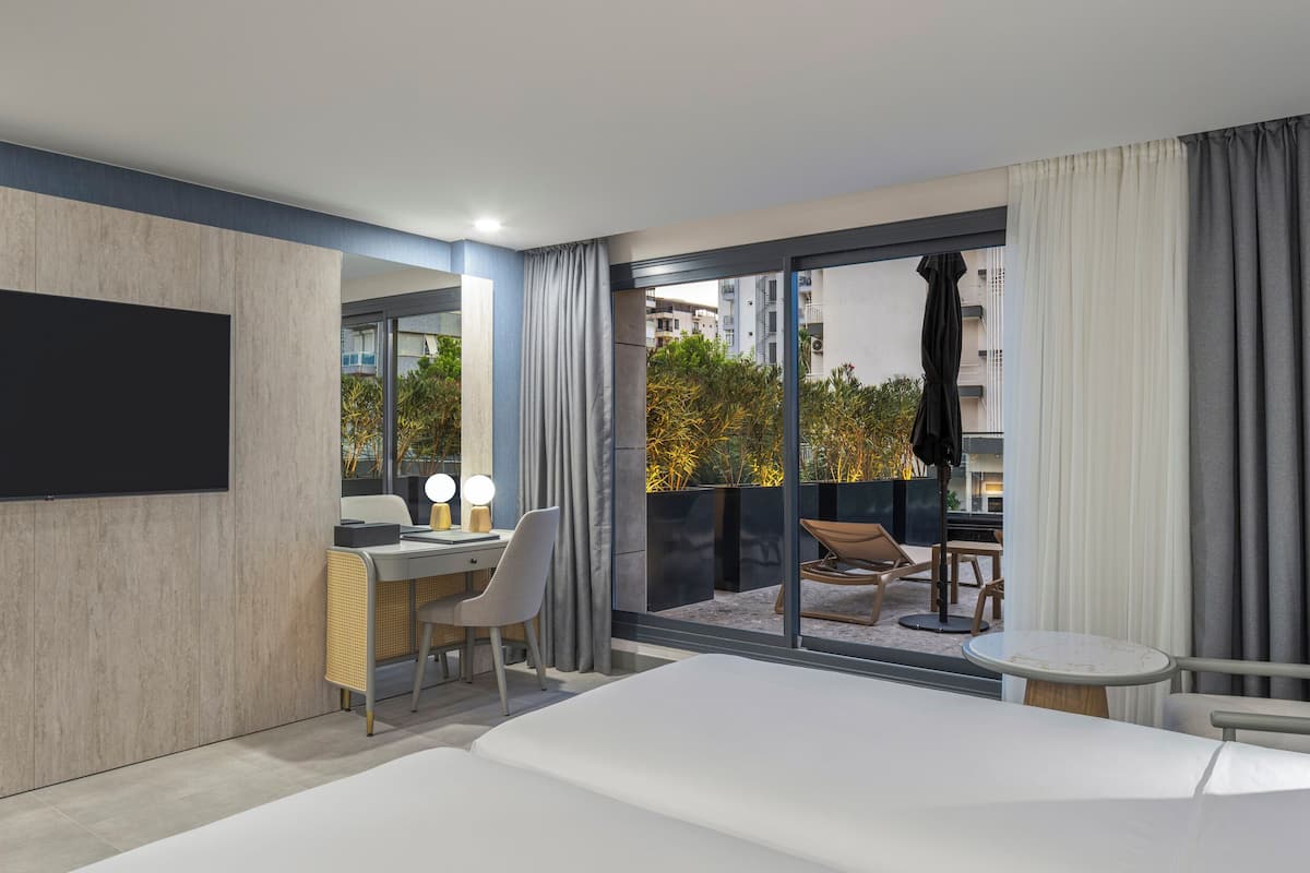 Una habitación de hotel moderna con una cama grande, un escritorio con una silla, una mesita pequeña y un balcón con vistas a la ciudad.