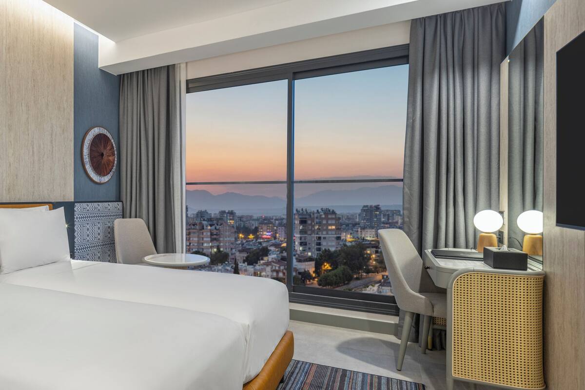 Habitación de hotel con una cama grande, un escritorio, una silla, una mesa redonda y vistas a un paisaje urbano al atardecer.