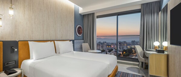 Une chambre d’hôtel avec un grand lit, un bureau, une chaise, une table ronde et une vue sur le paysage urbain au coucher du soleil.