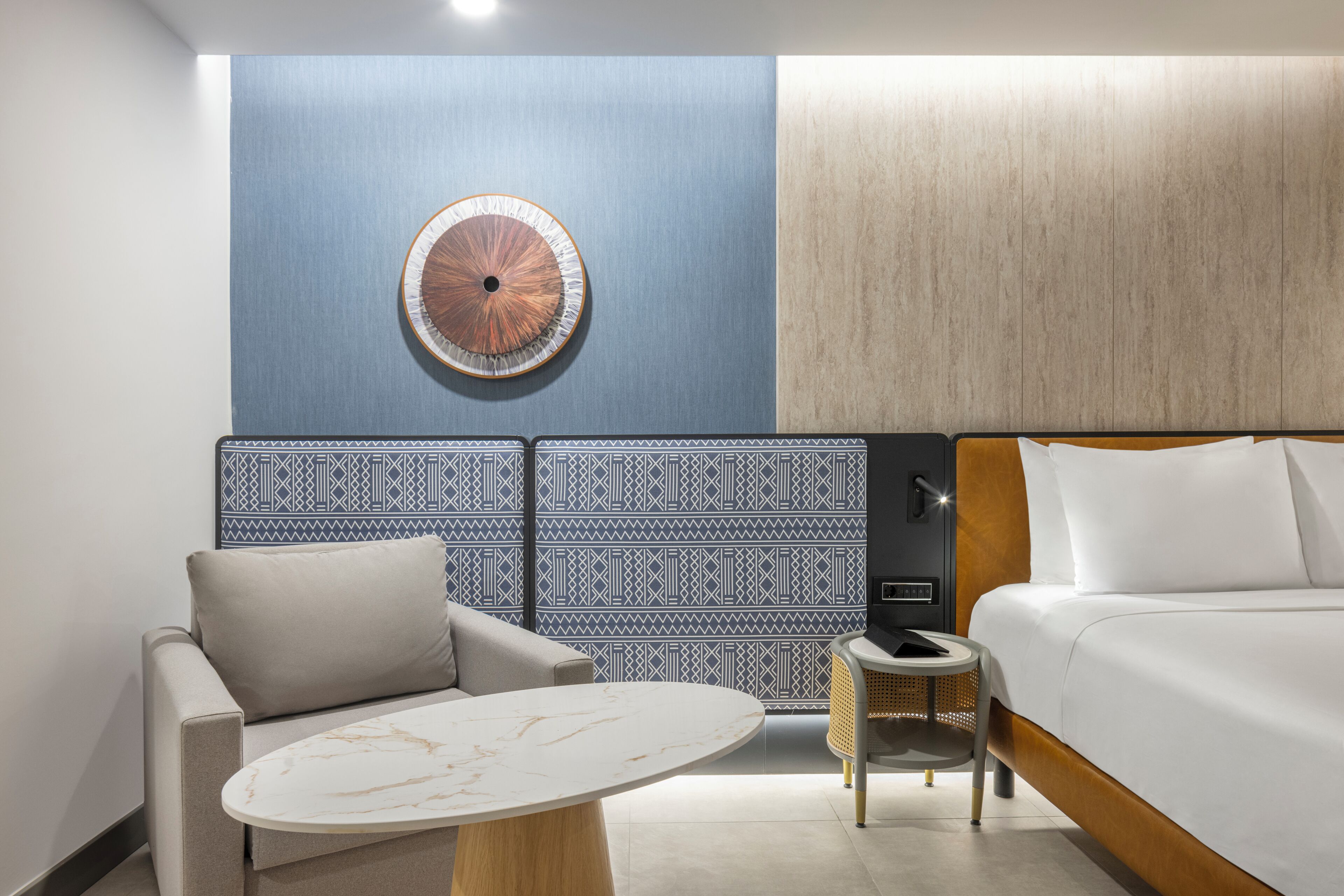 A hotel room with a bed, a round wall decoration, a patterned wall, a sofa, a small round table, and a nightstand.