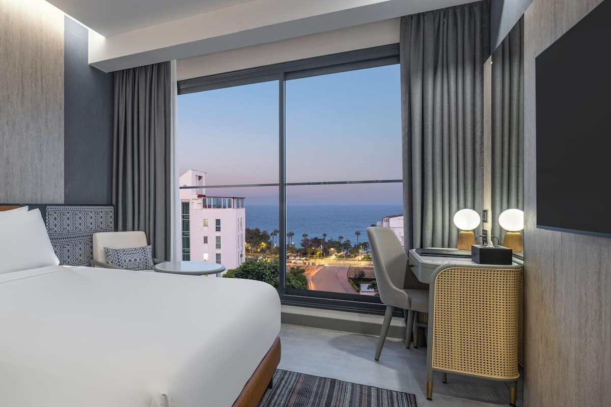 Una habitación de hotel moderna con una cama grande, un escritorio, una silla y vistas a la ciudad y al mar.