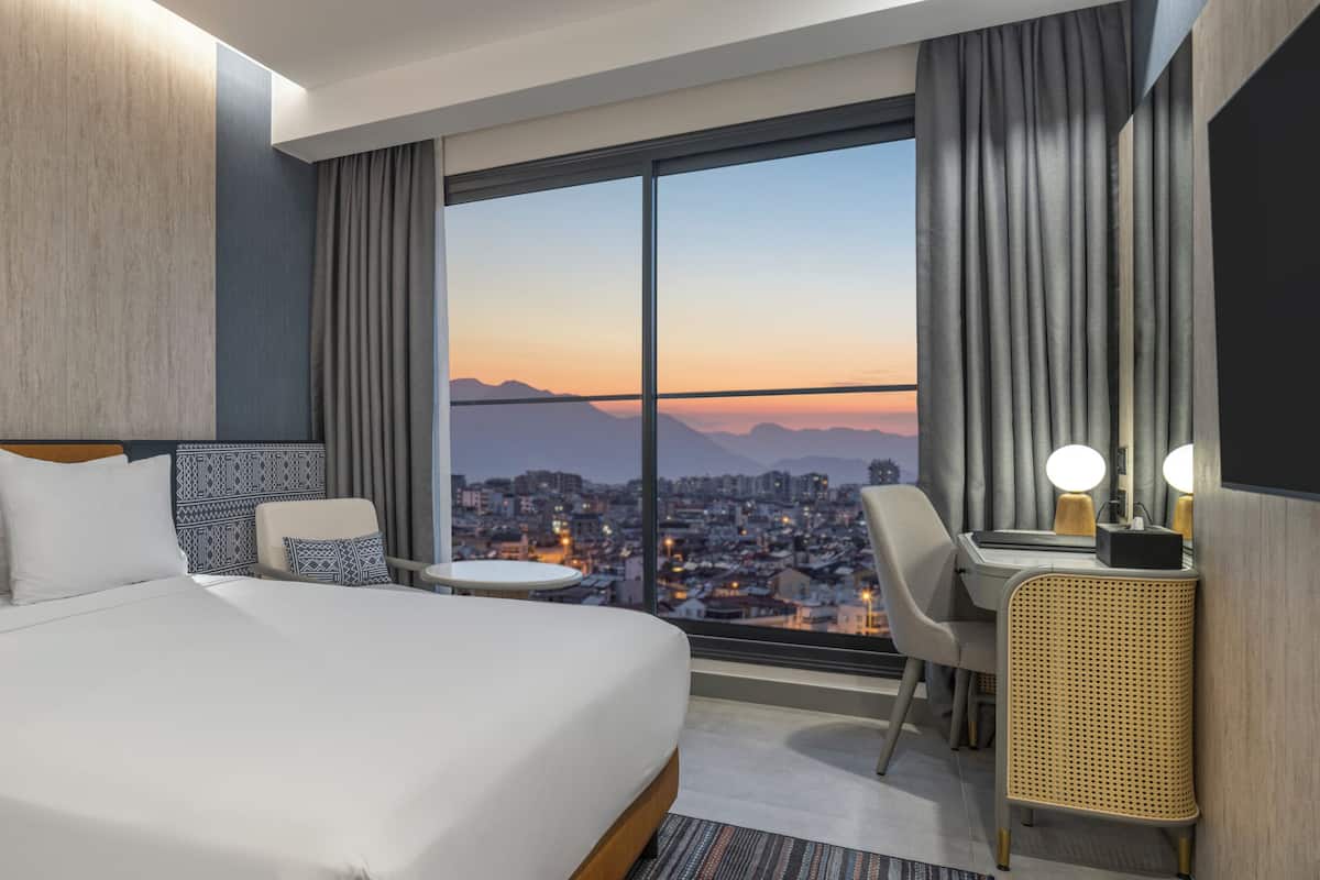 Habitación de hotel con una cama grande, un escritorio con lámpara, una silla, una mesita y vistas a un paisaje urbano al atardecer.