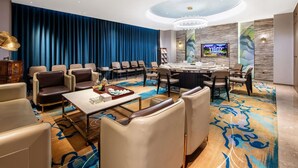 Hotelkamer met een ronde eettafel, meerdere stoelen, een groot blauw gordijn, een kroonluchter en een aan de muur bevestigde televisie.