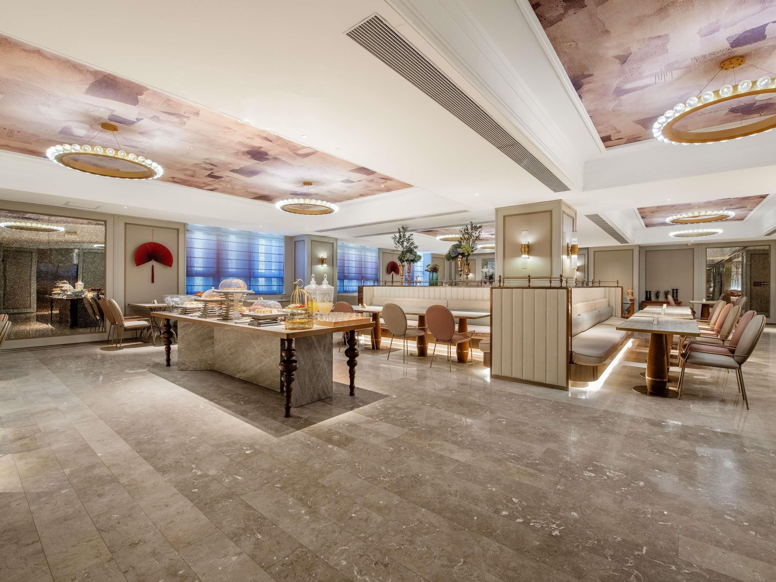 A spacious hotel lobby with a central dining area, seating arrangements, and a decorative ceiling.