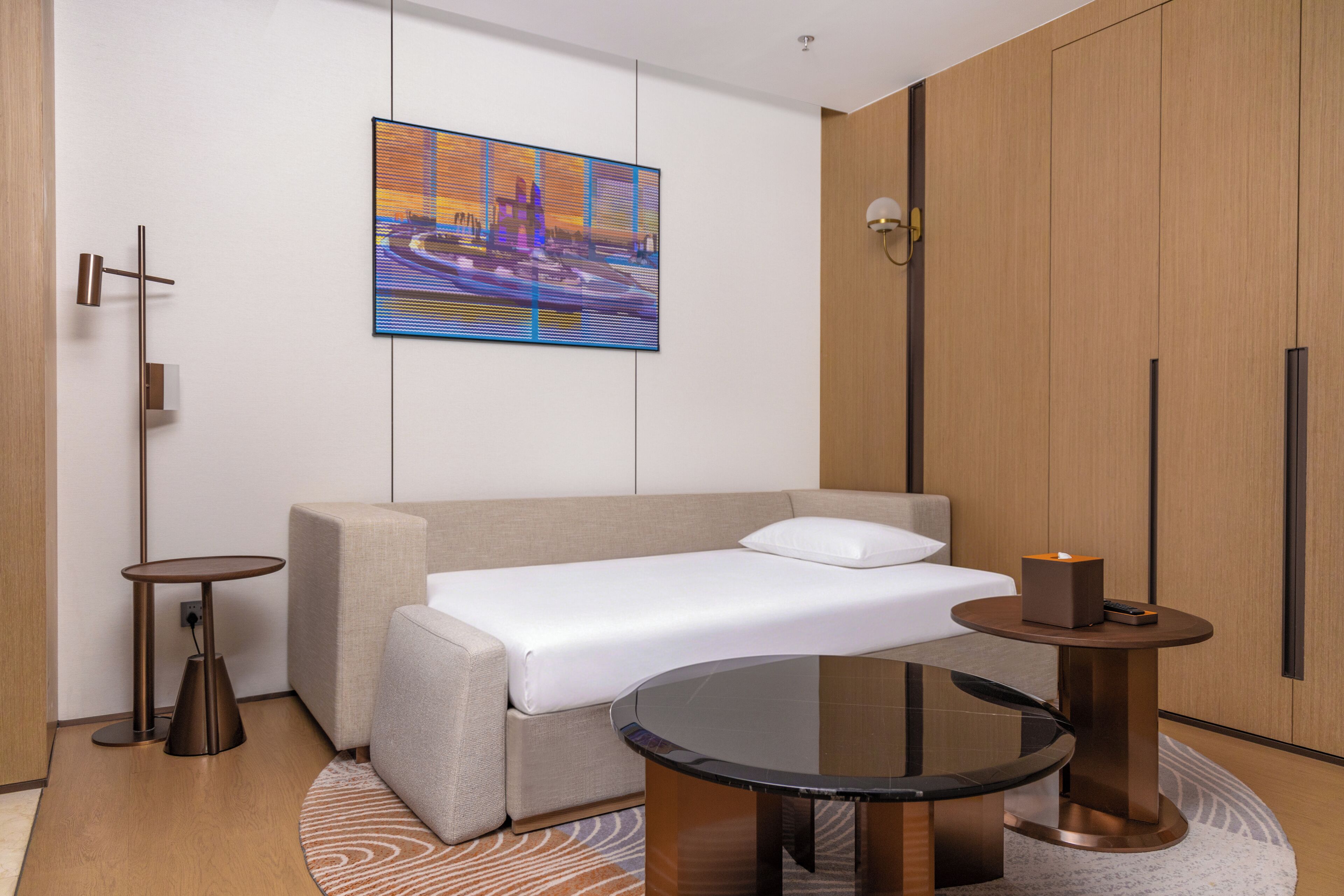 Una habitación de hotel moderna con sofá cama, una mesita redonda de vidrio, una pequeña mesita auxiliar y un cuadro en la pared.