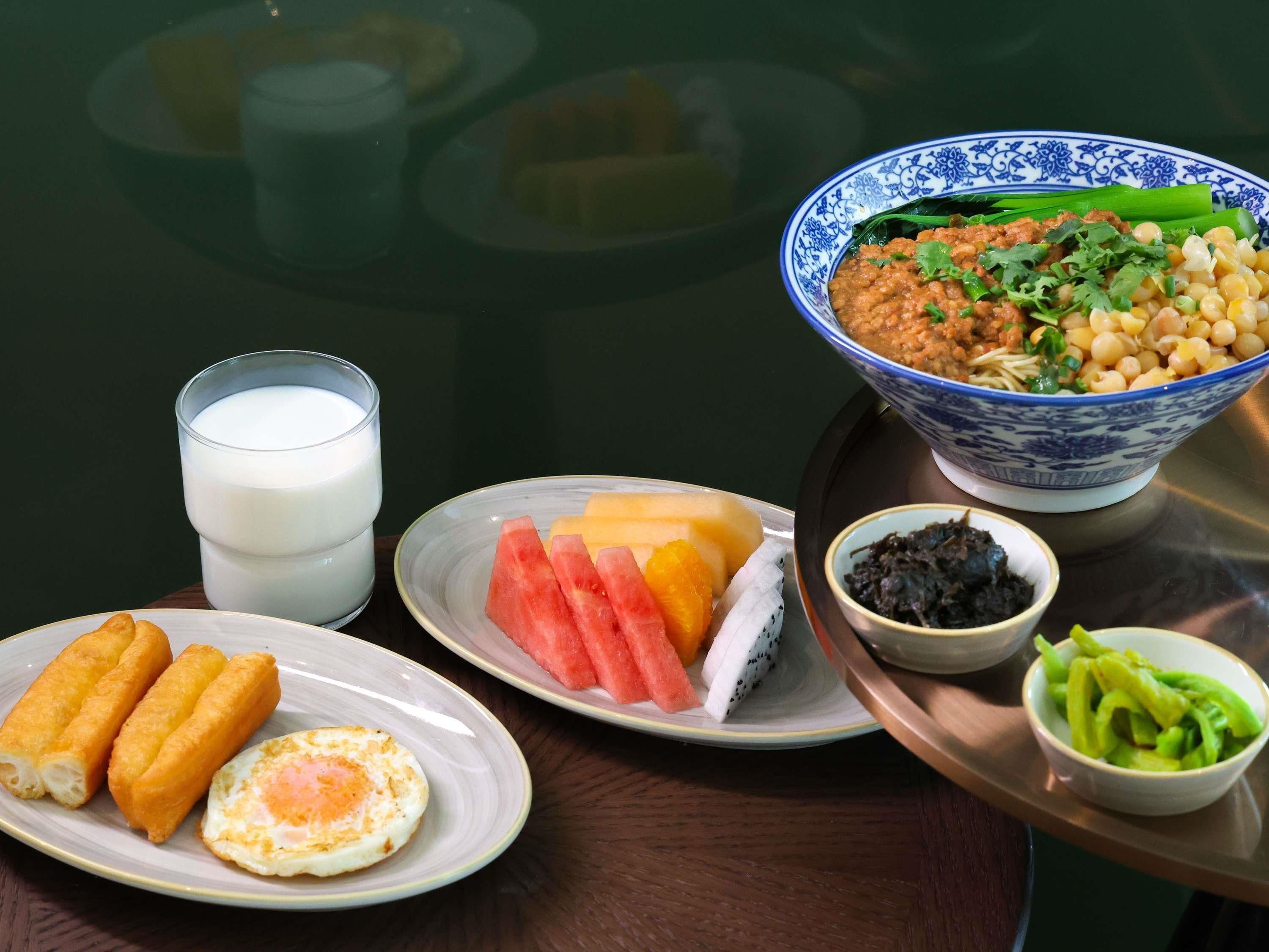 A table with a bowl of soup, a glass of milk, a plate with eggs and corn, a plate with fruit, and a bowl of vegetables.