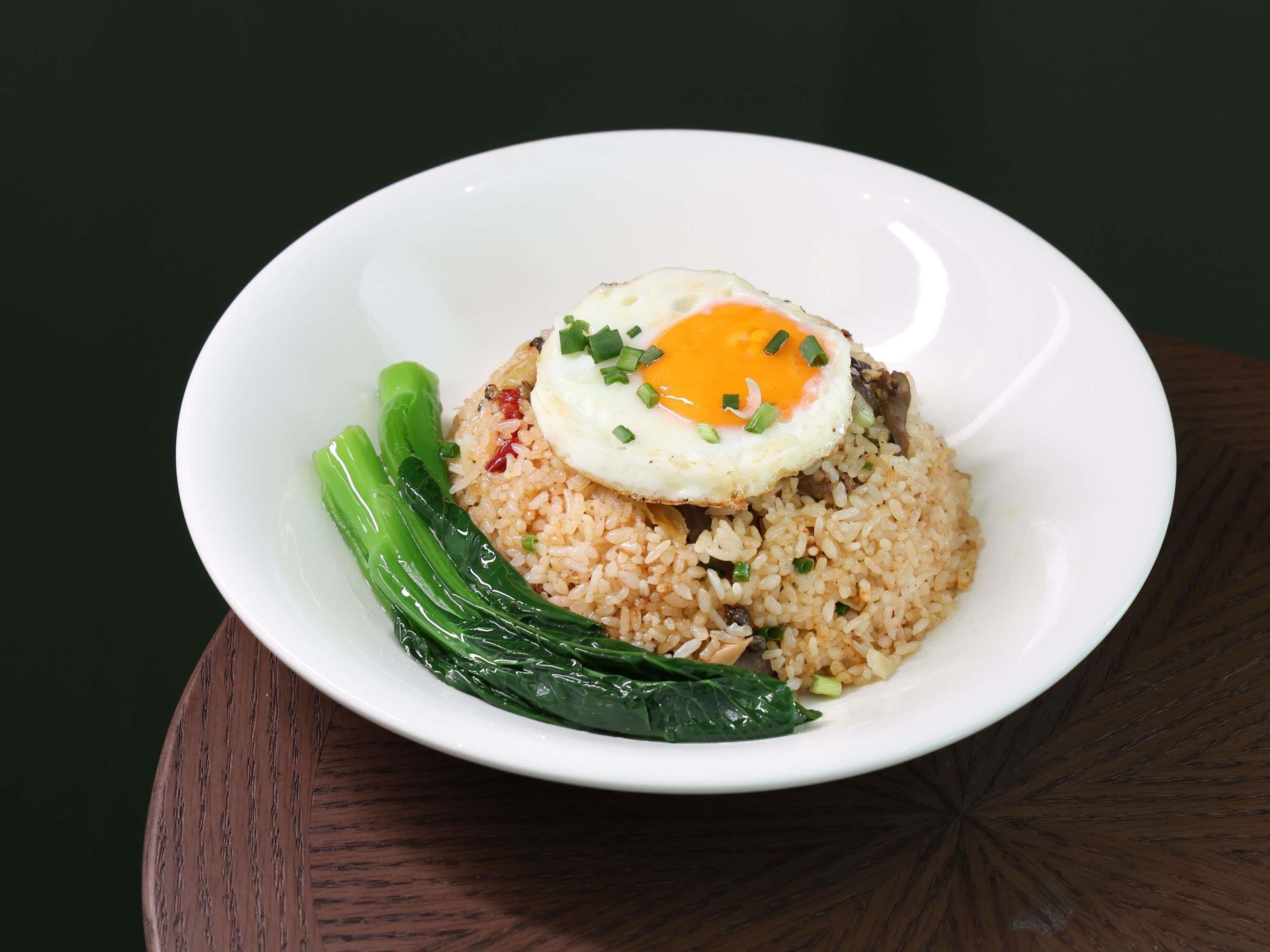 A white plate with fried rice, a fried egg, and green vegetables on a dark surface.
